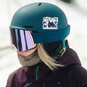 Pode incluir: Um capacete de esqui verde-azulado com um adesivo com um gr&aacute;fico de tr&ecirc;s mulheres e o texto "Girls Will Be Girls". O capacete tem uma al&ccedil;a azul escura e &oacute;culos espelhados com arma&ccedil;&atilde;o rosa. A pessoa est&aacute; usando uma jaqueta roxa escura.