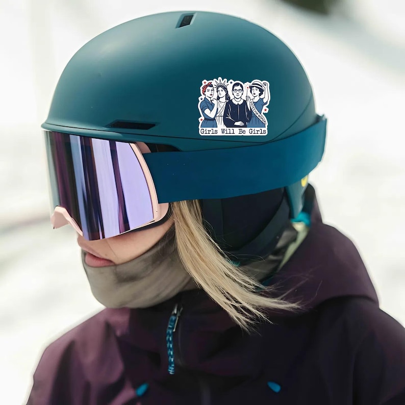 Pode incluir: Um capacete de esqui verde-azulado com um adesivo com um gr&aacute;fico de tr&ecirc;s mulheres e o texto "Girls Will Be Girls". O capacete tem uma al&ccedil;a azul escura e &oacute;culos espelhados com arma&ccedil;&atilde;o rosa. A pessoa est&aacute; usando uma jaqueta roxa escura.