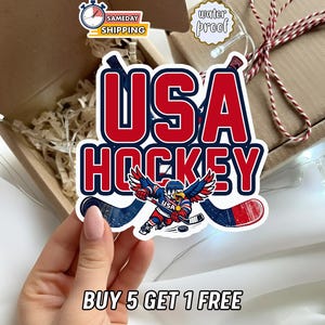 May include: A sticker with the words "USA HOCKEY" in red and blue. The sticker features an eagle in a hockey uniform with crossed hockey sticks. The text "BUY 5 GET 1 FREE" is at the bottom.