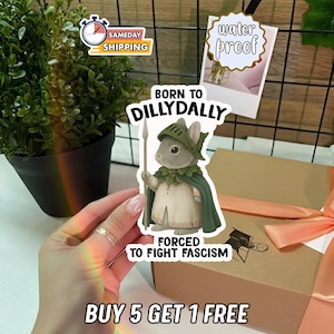 May include: A sticker featuring a cartoon rabbit in a helmet and cape, holding a spear. Text on the sticker reads "BORN TO DILLYDALLY" and "FORCED TO FIGHT FASCISM." Additional text says "BUY 5 GET 1 FREE" and "SAMEDAY SHIPPING."