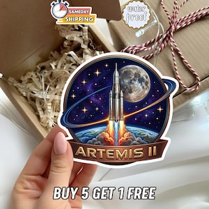 May include: A circular sticker featuring a space shuttle launching from Earth, with a full moon and starry background. The sticker has the text "ARTEMIS II" and "BUY 5 GET 1 FREE". The sticker is water proof.