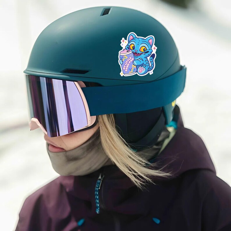 K&ouml;nnte beinhalten: T&uuml;rkisfarbener Skihelm mit einem Aufkleber einer blauen Cartoon-Katze, die eine Limonadendose mit der Aufschrift "My Little Soda Pop" h&auml;lt. Der Helm hat einen dunkelblauen Riemen und verspiegelte Brillen mit rosa Rahmen.