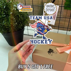Pode incluir: Um autocolante com um pato de desenho animado com o uniforme de hóquei da equipa dos EUA, segurando um stick e um disco de hóquei. O autocolante diz "TEAM USA HOCKEY" e "water proof". Uma caixa de presente com uma fita cor de pêssego está no fundo. O texto "BUY 5 GET 1 FREE" também é visível.