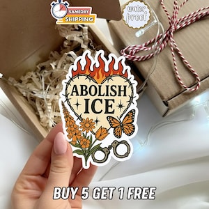 May include: A sticker with the text "ABOLISH ICE" inside a heart-shaped design with barbed wire, flames, flowers, and a butterfly. The sticker also features handcuffs and the phrase "BUY 5 GET 1 FREE".