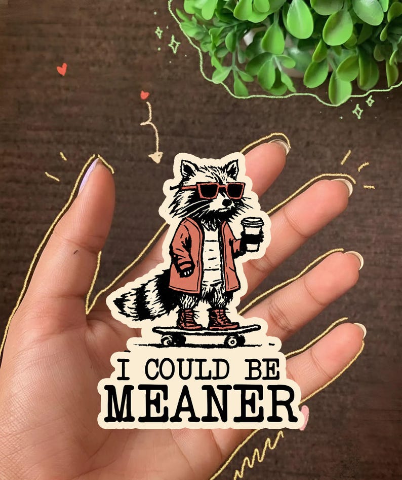 K&ouml;nnte beinhalten: Ein Aufkleber mit einem Waschb&auml;ren mit Sonnenbrille, roter Jacke und Stiefeln, der einen Kaffeebecher h&auml;lt und auf einem Skateboard steht. Der Aufkleber hat den Text "I COULD BE MEANER" in fetter Schrift.