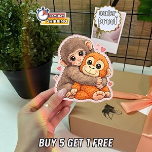Pode incluir: Um autocolante com dois macacos de desenho animado a abraçar-se, um castanho e outro laranja, com olhos grandes e corações. O autocolante está rotulado como "water proof" e o texto "BUY 5 GET 1 FREE" está na parte inferior.