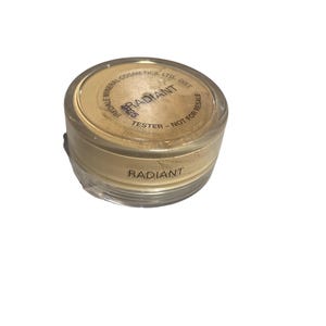 May include: A clear, cylindrical container of Radiant face powder. The lid has the word "RADIANT" printed on it. The container is labeled "TESTER - NOT FOR RESALE". The powder is a light beige color.