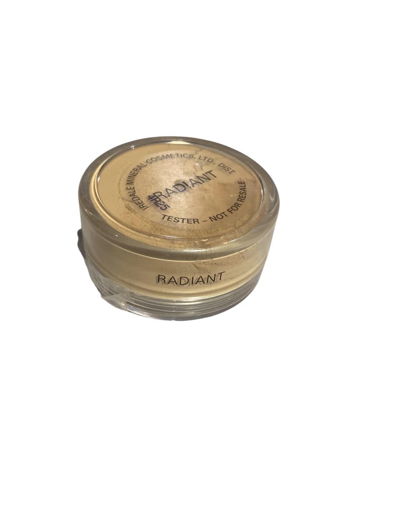 May include: A clear, cylindrical container of Radiant face powder. The lid has the word "RADIANT" printed on it. The container is labeled "TESTER - NOT FOR RESALE". The powder is a light beige color.