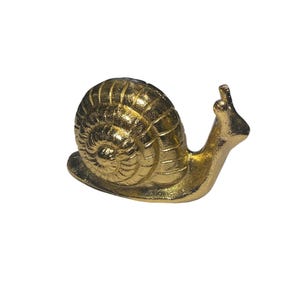 May include: A golden snail figurine with a textured shell and detailed body. The snail is posed on a flat surface, with its antennae raised. The figurine is made of a shiny, metallic material.