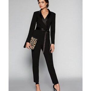 May include: A black jumpsuit with a satin lapel and a self-tie belt. The jumpsuit has long sleeves and a fitted silhouette. The model is wearing black heels and holding a leopard print clutch.