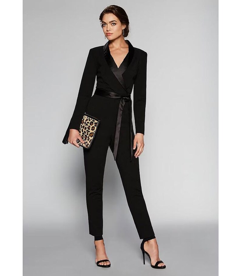 May include: A black jumpsuit with a satin lapel and a self-tie belt. The jumpsuit has long sleeves and a fitted silhouette. The model is wearing black heels and holding a leopard print clutch.