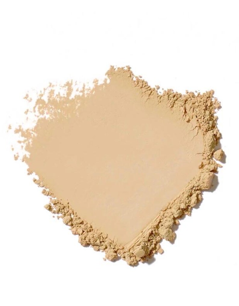May include: A close-up of a light beige powder product, possibly a cosmetic, against a white background. The powder is finely milled, with a smooth, even surface and a slightly textured edge. The shape is irregular, with a rectangular form.