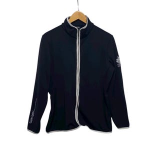 Może przedstawiać: Czarna kurtka zapinana na zamek z wysokim kołnierzem i białym wykończeniem. Kurtka ma biały zamek błyskawiczny i napis "GALVIN GREEN" na lewym rękawie. Logo znajduje się na prawym rękawie. Kurtka wisi na drewnianym wieszaku.