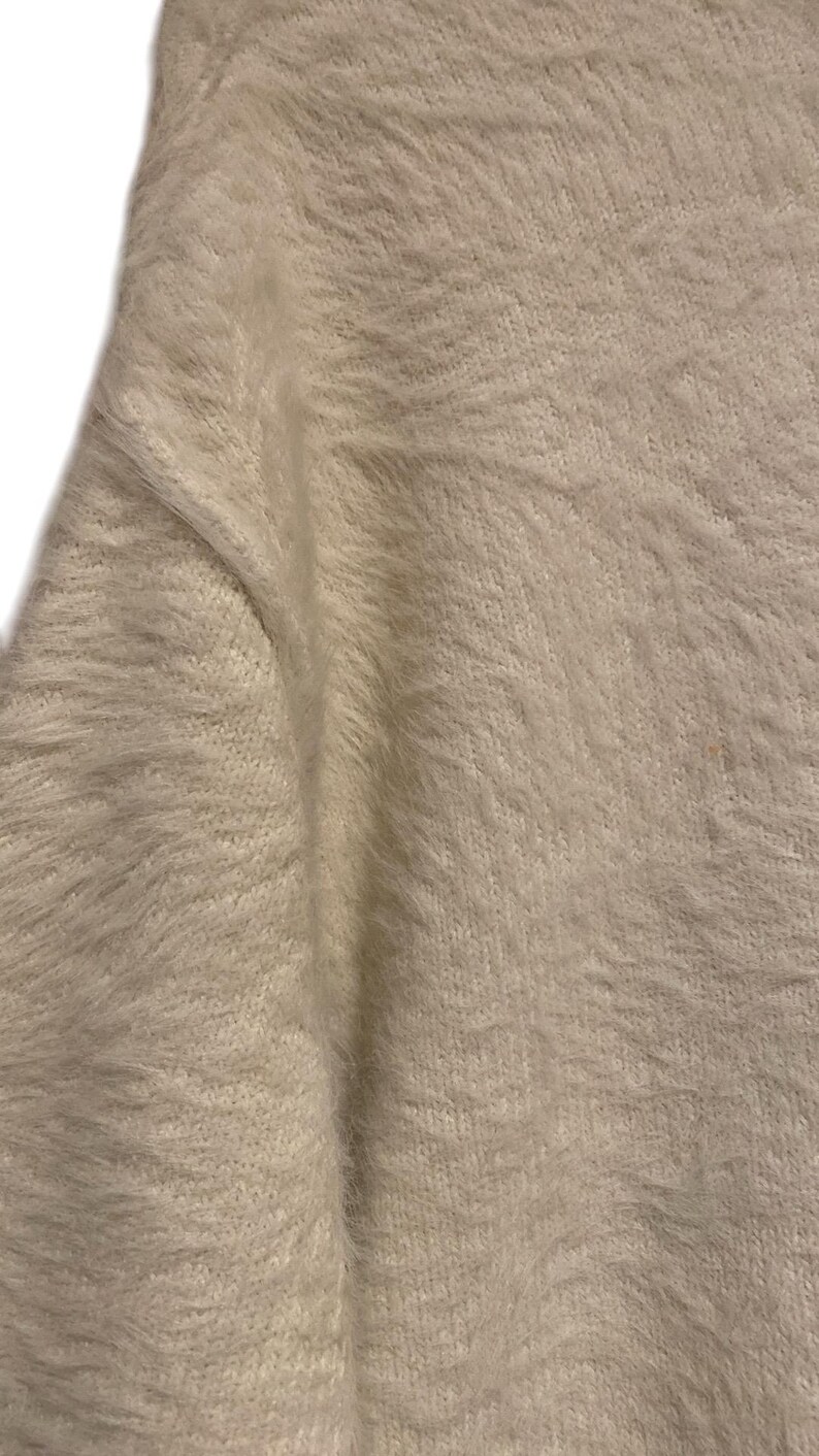 May include: Close-up of a white fuzzy sweater with a soft, textured knit.