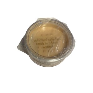 May include: A round, clear plastic container with a beige powder inside, sealed with clear plastic wrap. The text "puncture selected holes to control volume" is visible on the lid. The container is on a white background.