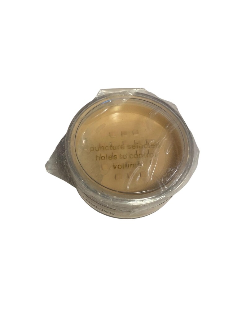 May include: A round, clear plastic container with a beige powder inside, sealed with clear plastic wrap. The text "puncture selected holes to control volume" is visible on the lid. The container is on a white background.