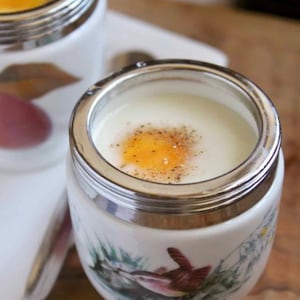 May include: Close-up of a white porcelain egg cup with a silver rim, filled with a cooked egg and sprinkled with pepper. The cup features a painted bird and floral design. The egg yolk is bright yellow.