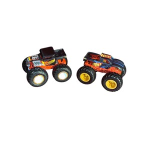 May include: Two Hot Wheels monster trucks. One truck is black and orange with a skull design and the words "Hot Wheels". The other truck is black and red with the words "Hot Wheels". Both trucks have large black tires with gold rims.