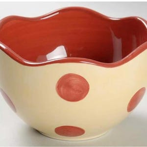 May include: A ceramic bowl with a cream-colored exterior and a red interior. The bowl has a wavy rim and is decorated with large red polka dots. This decorative bowl is suitable for serving food.