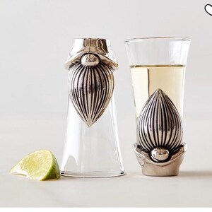 May include: Two clear shot glasses with silver gnome-shaped stoppers, one filled with a light-colored liquid. A lime wedge sits nearby. The stoppers have detailed beard designs. The glasses are on a white surface.