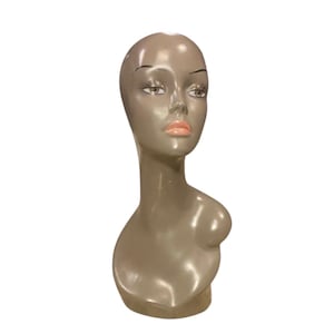 May include: A mannequin head and bust in a neutral taupe color. The face has painted features including dark eyebrows, eyelashes, and pink lips. The bust is smooth and glossy, with a rounded shoulder.