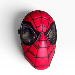 May include: A red and black Spider-Man mask featuring a prominent web pattern across the surface. The mask includes two large, dark, translucent eye lenses with a subtle zigzag texture visible within the lens material and a black frame border.