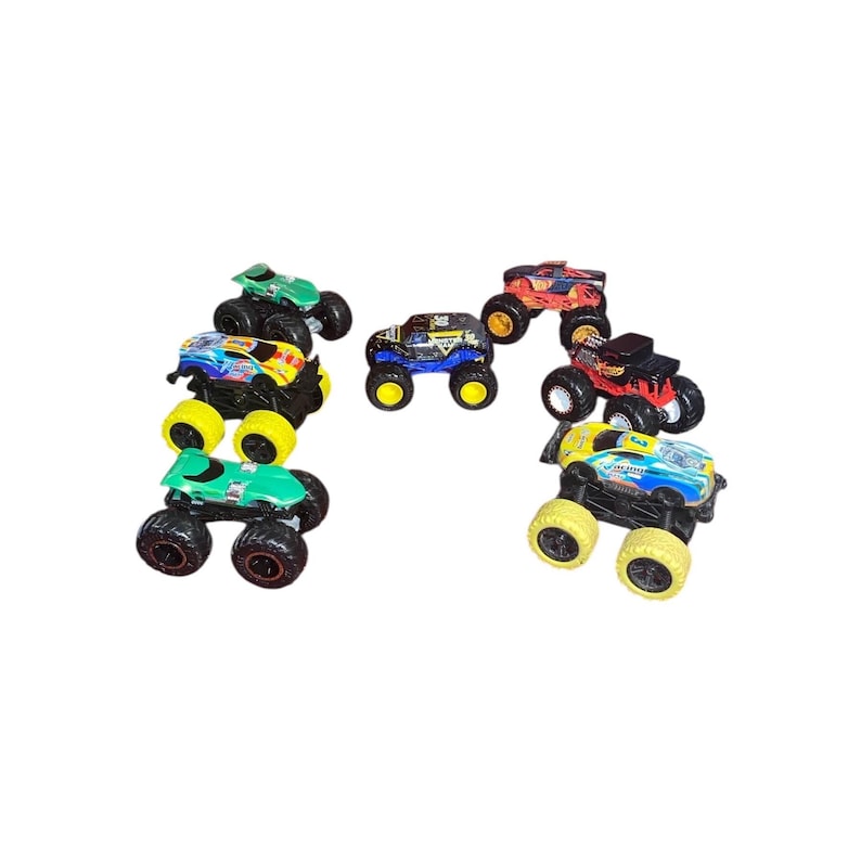 May include: A collection of toy monster trucks in various colors, including green, blue, red, and yellow. Each truck features large, oversized wheels and detailed designs. The trucks are arranged in a semi-circular pattern against a white background.