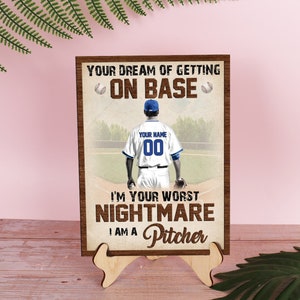 Puede incluir: Un letrero de madera con un jugador de béisbol en un campo. El letrero dice "Your dream of getting on base I'm your worst nightmare I am a pitcher".