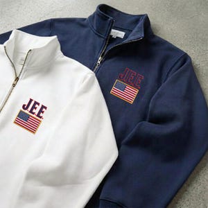 Könnte beinhalten: Zwei Sweatshirts mit Reißverschluss, eines weiß und eines marineblau. Jedes zeigt den Text "J.E.E." über einem gestickten amerikanischen Flaggenaufnäher. Die Sweatshirts haben einen Stehkragen und einen goldfarbenen Reißverschluss.