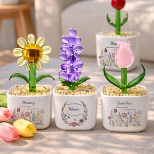 May include: Glass flower sculptures in white pots with the words "Mommy", "Mama", and "Grandma". The flowers are a sunflower, hyacinth, and tulip. Each pot has a floral design and the phrase "love is the flower, you are the seed."