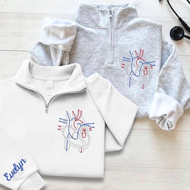 Puede incluir: Dos sudaderas con cremallera de un cuarto, una blanca y otra gris, cada una bordada con un dise&ntilde;o detallado de coraz&oacute;n anat&oacute;mico en hilo rojo, azul y gris. La sudadera blanca tiene el nombre "Evelyn" bordado en la manga.