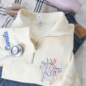 Puede incluir: Sudadera crema con cremallera de cuarto de cremallera y un diagrama del coraz&oacute;n en rojo y azul. El nombre "Camila" est&aacute; impreso en azul en la manga izquierda. Un estetoscopio descansa sobre la sudadera. La sudadera est&aacute; sobre unos vaqueros.