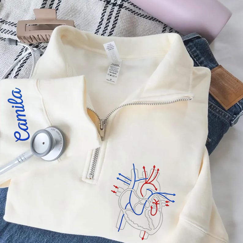 Puede incluir: Sudadera crema con cremallera de cuarto de cremallera y un diagrama del coraz&oacute;n en rojo y azul. El nombre "Camila" est&aacute; impreso en azul en la manga izquierda. Un estetoscopio descansa sobre la sudadera. La sudadera est&aacute; sobre unos vaqueros.