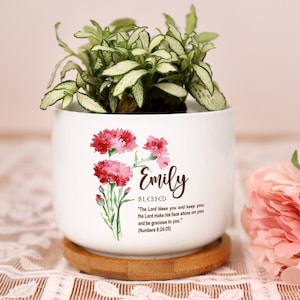 May include: A white ceramic plant pot featuring a floral design and the name "Emily". The pot contains a small plant with green and white leaves. It rests on a round, wooden saucer. Text on the pot reads "Blessed" and a Bible verse.