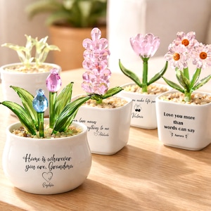 Puede incluir: Varios pequeños maceteros de cerámica blanca con esculturas de flores de cristal. Las flores son rosas, azules y transparentes, con tallos y hojas de cristal verde. Cada maceta tiene un mensaje.