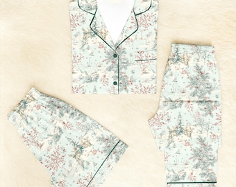 Könnte beinhalten: Hellblaues Pyjama-Set mit Winterlandschaftsdruck, einschließlich Bäumen, Hirschen und Schnee. Das Set besteht aus einem Hemd mit Knöpfen und grünen Paspeln, Shorts und einer langen Hose. Der Pyjama ist aus weichem Stoff.