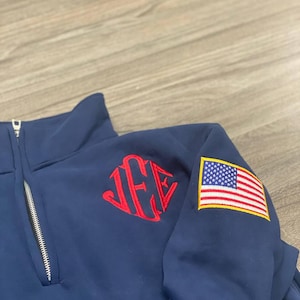 May include: A navy blue quarter-zip pullover featuring a red embroidered monogram on the chest and a rectangular American flag patch on the sleeve. The garment has a silver-toned metal zipper pull and a high collar design with visible stitching.