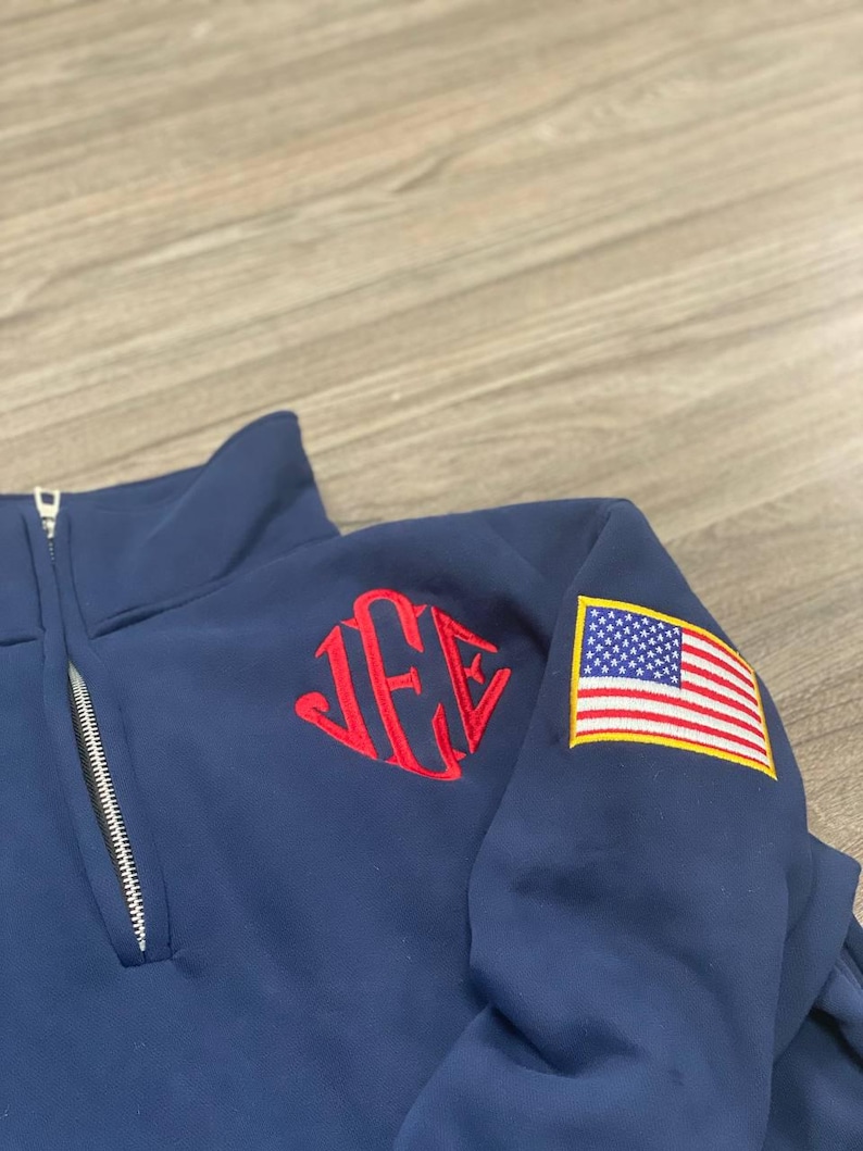 May include: A navy blue quarter-zip pullover featuring a red embroidered monogram on the chest and a rectangular American flag patch on the sleeve. The garment has a silver-toned metal zipper pull and a high collar design with visible stitching.