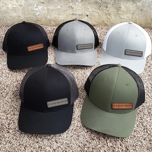 Puede incluir: Cinco gorras de camionero en negro, gris, blanco y verde oliva. Cada gorra tiene una parte trasera de malla y un panel frontal sólido con un parche de cuero que muestra números romanos. Las gorras están dispuestas sobre una superficie texturizada.