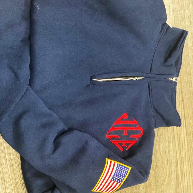 May include: A navy blue quarter-zip pullover sweatshirt featuring a red embroidered monogram logo on the left chest and a small rectangular American flag patch stitched onto the lower left sleeve near the cuff.