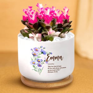 May include: A white ceramic plant pot with a vibrant pink and green succulent plant. The pot features a floral design with the name "Emma" and the word "HOPE" along with a Bible verse. The pot sits on a round wooden base.