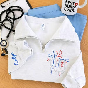 May include: A light grey quarter-zip sweatshirt featuring a detailed embroidered heart diagram in red and blue. The sleeve is embroidered with the name "Evelyn" in blue. A white mug with "BEST NURSE EVER" is visible.