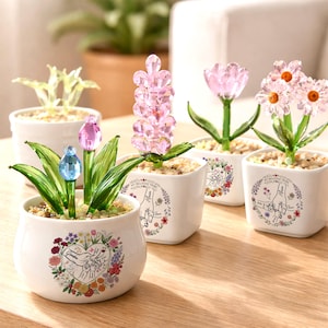 May include: A collection of decorative crystal flower arrangements in small white pots. The flowers include pink tulips, daisies, and hyacinths, with green glass leaves. The pots are decorated with floral designs and the words "My Beloved Mom."
