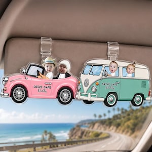 Puede incluir: Dos adornos para visera de coche. Uno es un descapotable rosa con dos bebés, el otro es una furgoneta turquesa y blanca con dos bebés. Ambos tienen el texto "DRIVE SAFE Daddy".
