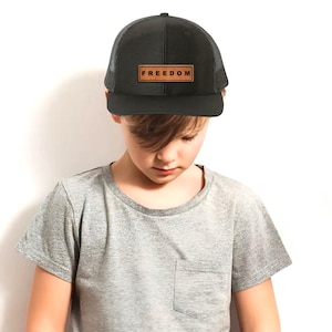 Könnte beinhalten: Schwarze Trucker-Cap mit Mesh-Rückseite und gebogenem Schirm. Die Kappe hat einen rechteckigen braunen Lederaufnäher mit dem Wort "FREEDOM". Die Person trägt ein graues T-Shirt.