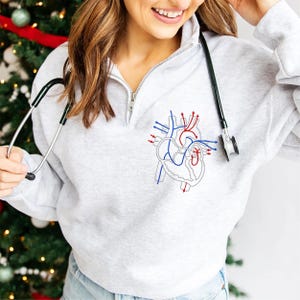 Puede incluir: Sudadera gris claro con cremallera de un cuarto de cuello, con un dise&ntilde;o de coraz&oacute;n anat&oacute;mico bordado en rojo y azul. Un estetoscopio cuelga alrededor del cuello. La sudadera tiene un cuello alto y una cremallera plateada.