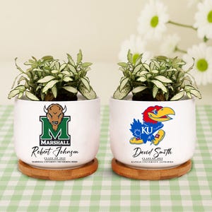 May include: Two white ceramic planters with green plants, each on a wooden coaster. One planter features a green and black Marshall University logo with the name "Robert Johnson" and "Class of 2025". The other displays a blue and red Kansas Jayhawks logo with the name "David Smith".