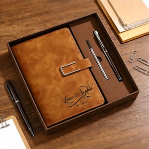 May include: A brown leather journal with a silver clasp, presented in a gift box with two pens. The journal is embossed with the names "Liam & Sophia" and the date "EST. 20.10.23". The set includes a pen and a ballpoint pen.