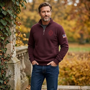 May include: A maroon quarter-zip pullover featuring a silver zipper, a dark red embroidered monogram on the left chest, and a small American flag patch on the upper left sleeve, paired with dark blue denim jeans.
