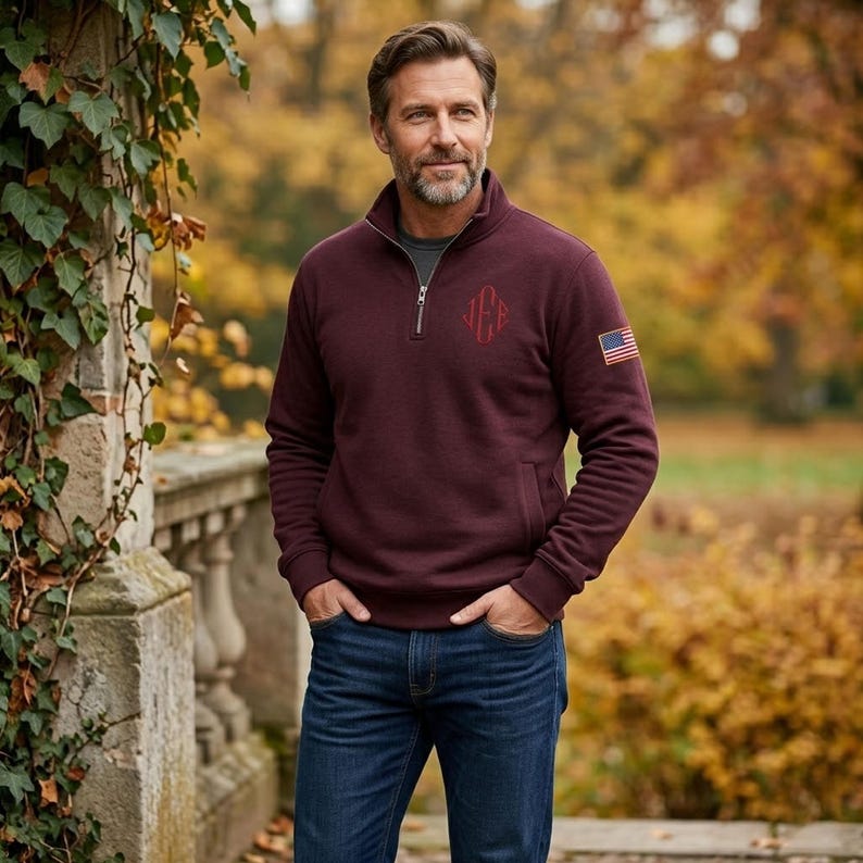 May include: A maroon quarter-zip pullover featuring a silver zipper, a dark red embroidered monogram on the left chest, and a small American flag patch on the upper left sleeve, paired with dark blue denim jeans.
